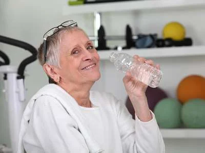 Ältere Frau mit kurzen grauen Haaren und Brille auf dem Kopf hält eine Wasserflasche und lächelt in einem Raum mit Sportgeräten und bunten Gymnastikbällen im Hintergrund.