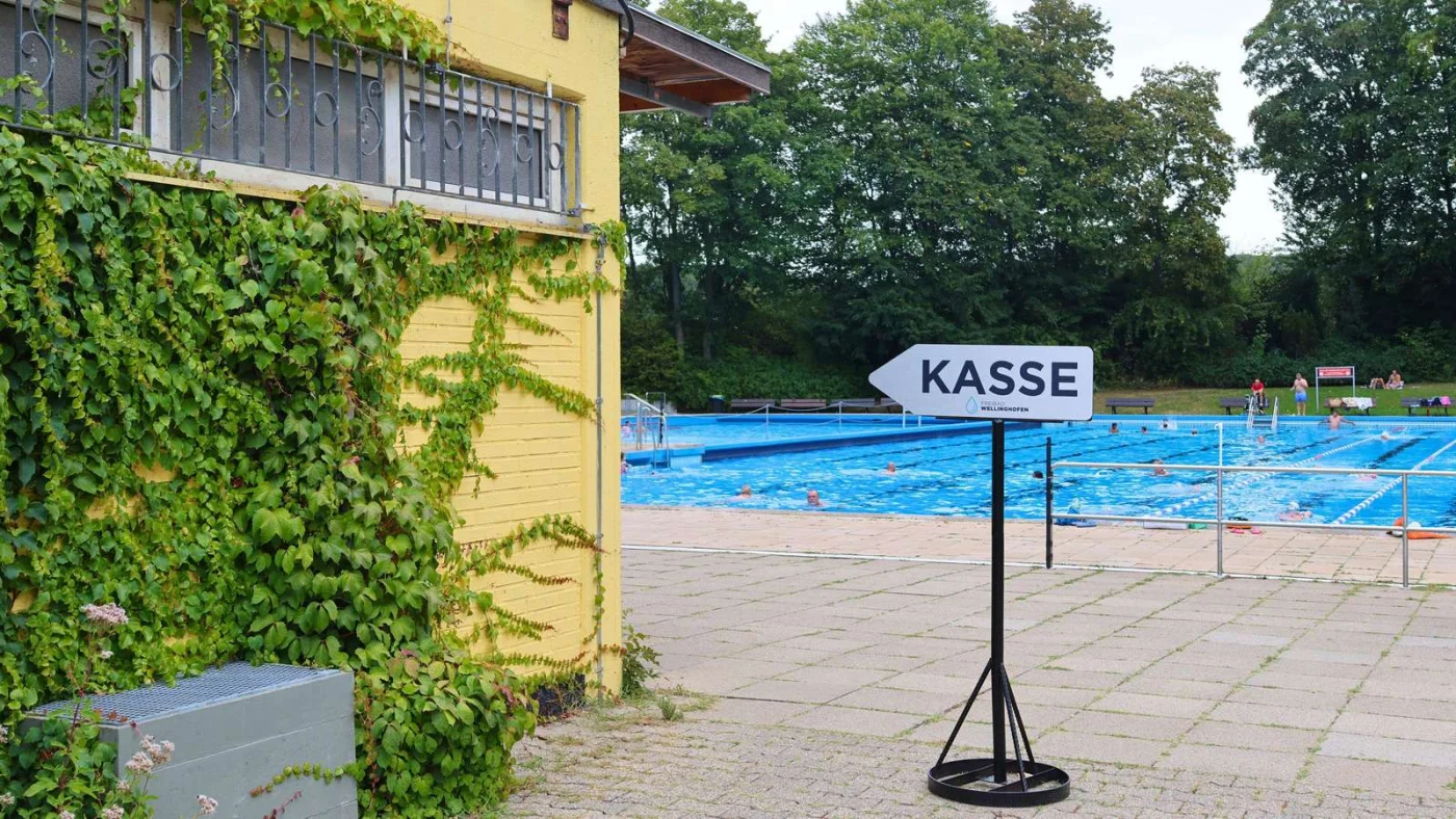 Blick auf das Schwimmerbecken und ein Schild zum Kassenhäuschen.