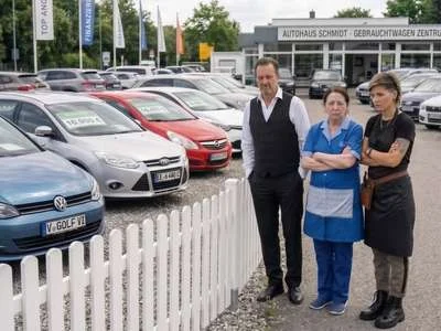 Drei Personen in Berufskleidung stehen vor einem Zaun, dahinter stehen Gebrauchtwagen.