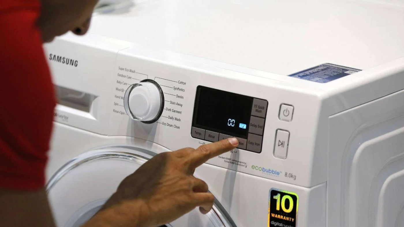 Eine Hand bedient das digitale Display einer weißen Samsung-Waschmaschine mit Drehknopf und Aufkleber „10 WARRANTY“.