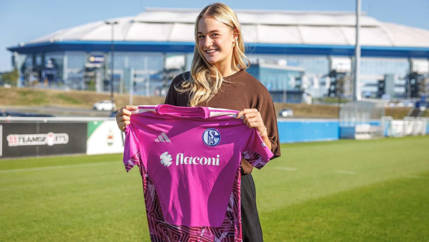 Josefine Osigus hält ein pinkes Schalker Torwarttrikot hoch. Im Hintergrund ist die Veltins-Arena.