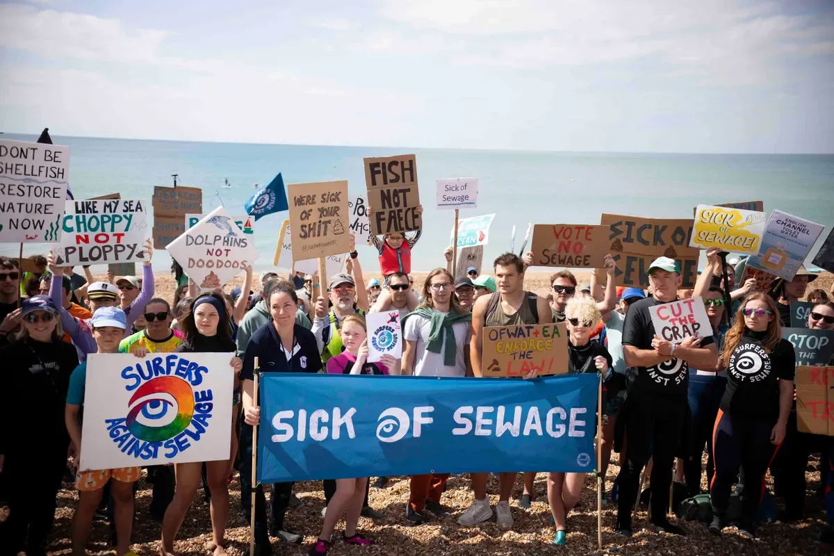 Surfers on a beach hold up signs including one that says: ‘Sick of Sewage’