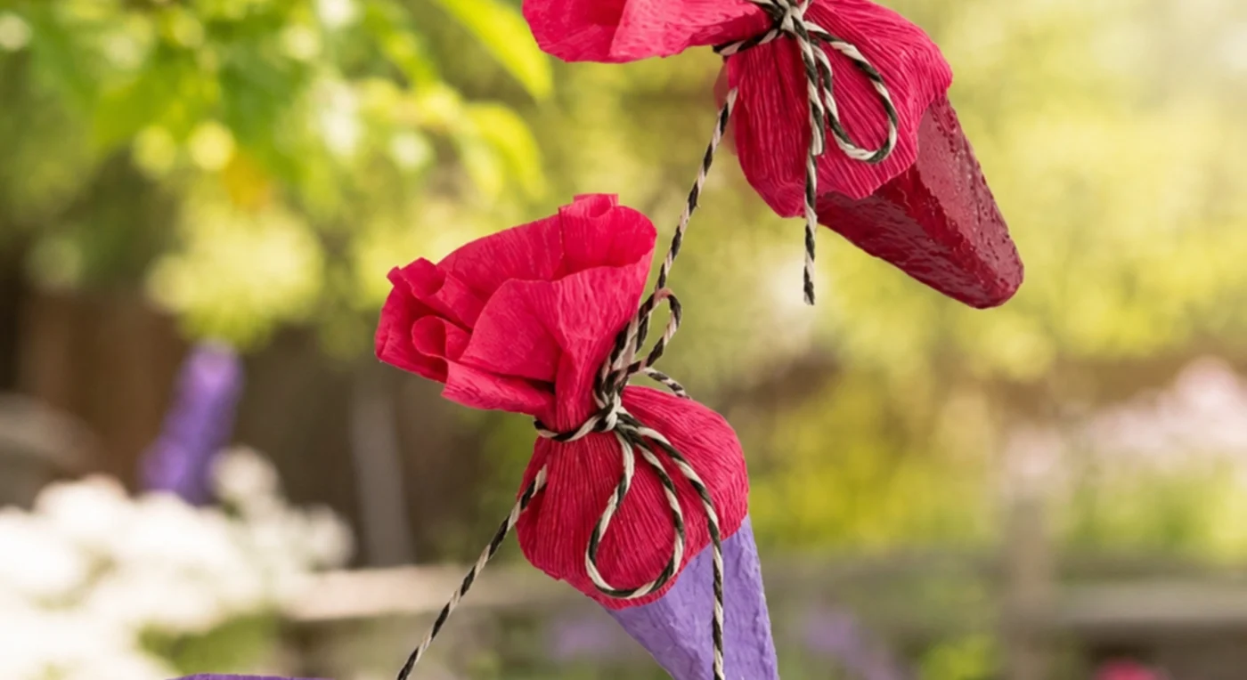 Vier bunte Mini-Schultüten aus Krepppapier und Eierkarton hängen an Kordeln vor einem sonnigen, unscharfen Gartenbeet mit Blumen und Bäumen.