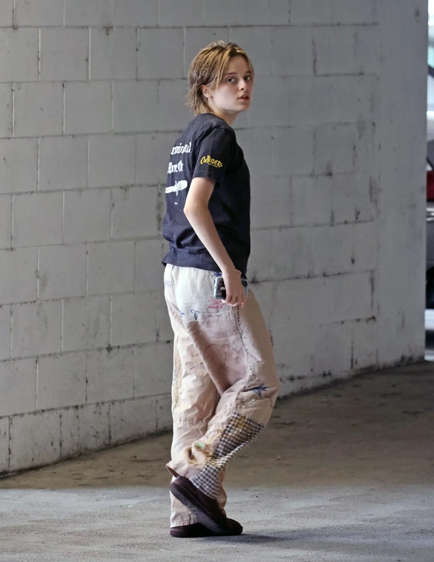 Vivienne Jolie walking away, turning her head to look back, carrying a cell phone in her hand.