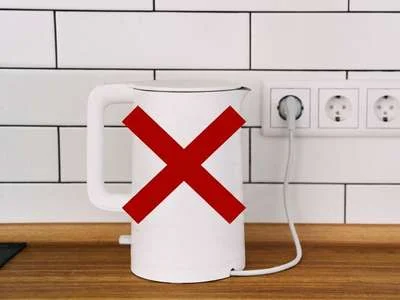 Weißer Wasserkocher auf Holzarbeitsplatte vor weißer Fliesenwand mit rotem X über dem Wasserkocher.
