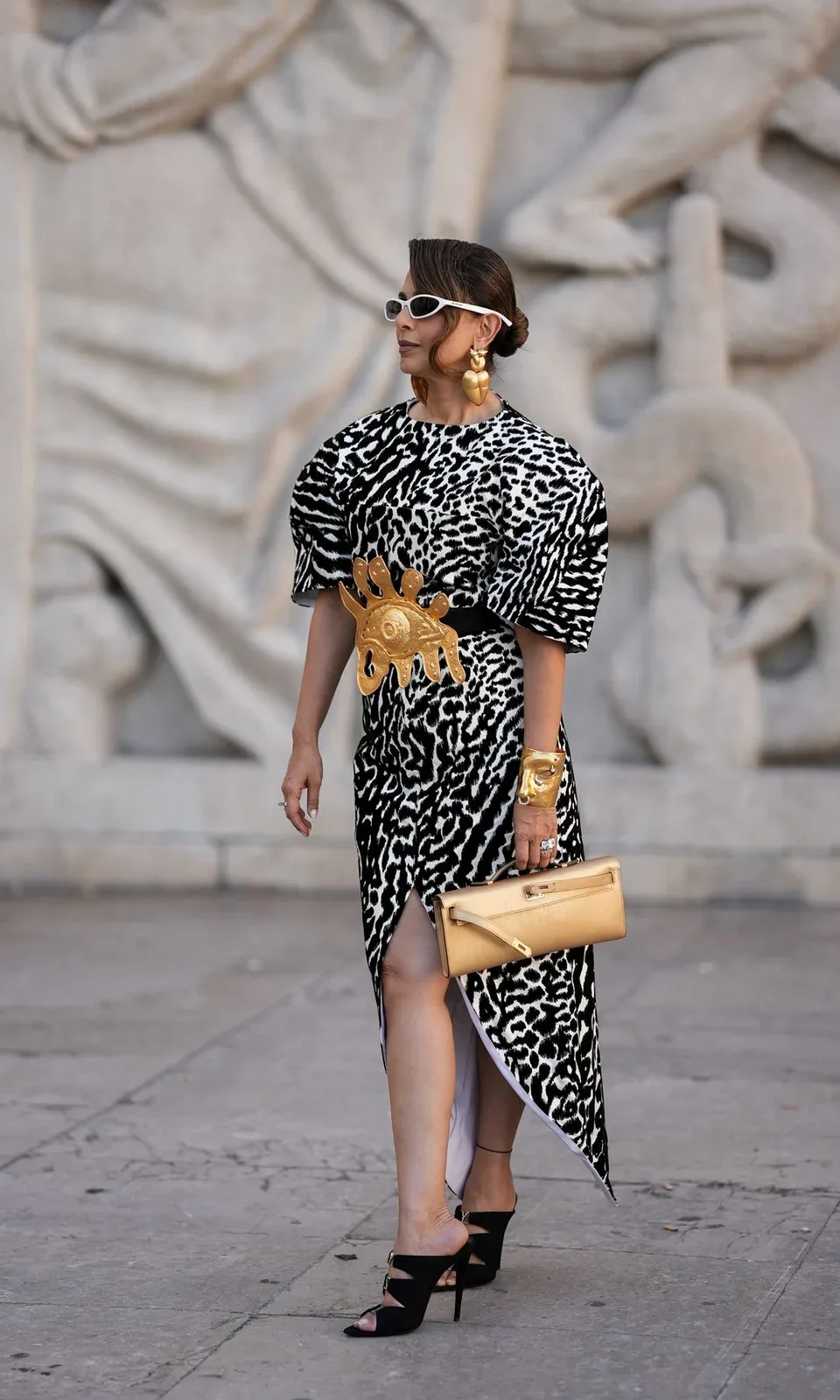 Woman in leopard print dress, gold accessories, and sunglasses stands on sidewalk.