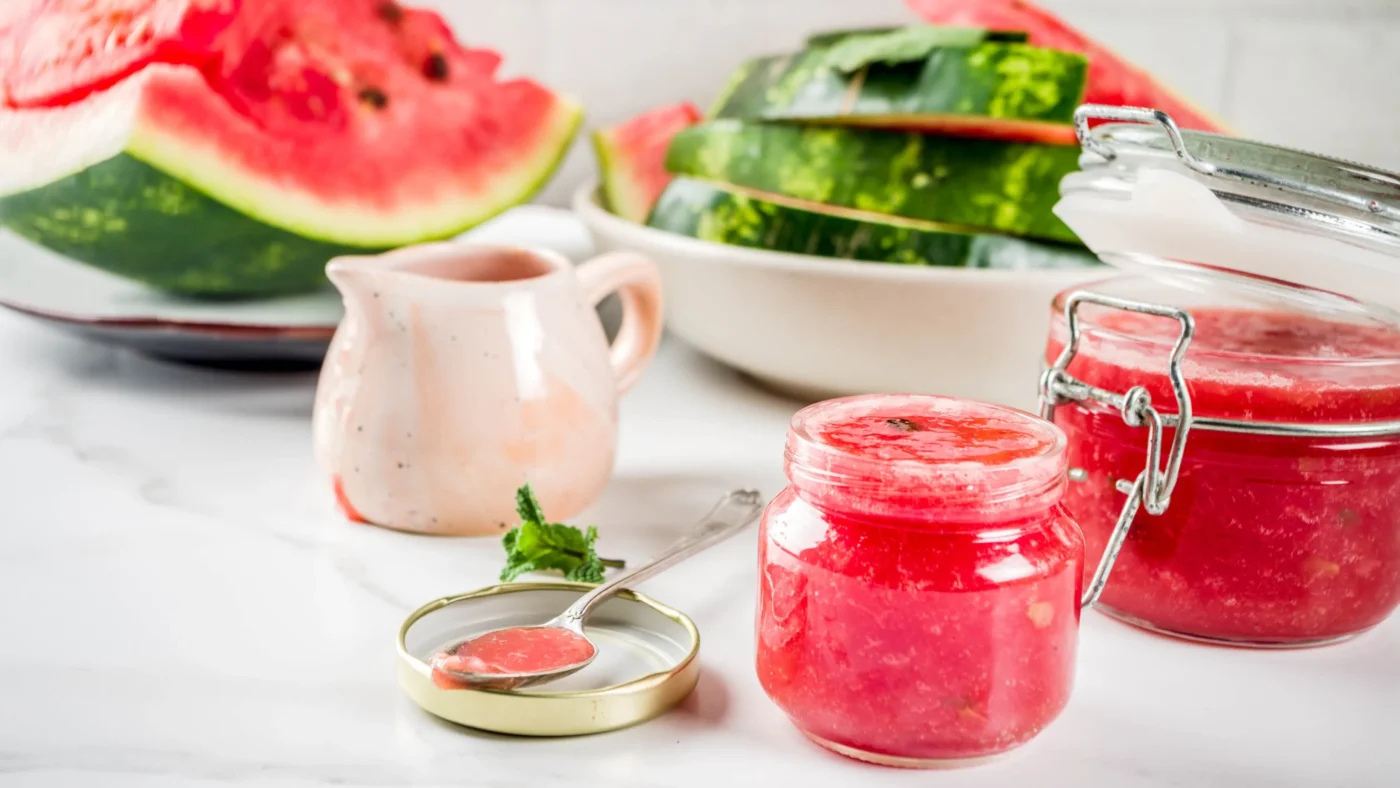 Zwei Gläser mit Wassermelonen-Marmelade stehen auf einer Marmorplatte, dahinter einige Wassermelonen-Stücke mit grüner Haut.