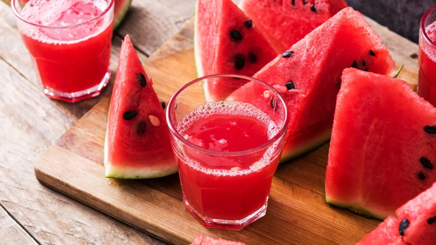 Zwei Gläser mit Wassermelonen-Smoothie stehen auf einer Holzplatte zwischen Wassermelonenstücken.