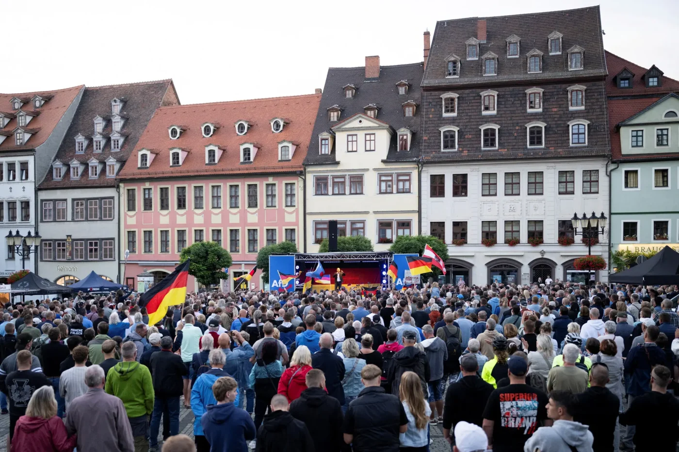 A crowd fills a town square, standing before a stage branded with the logo of the AfD. Pastel-colored buildings, with steep tiled roofs, rise behind the stage.