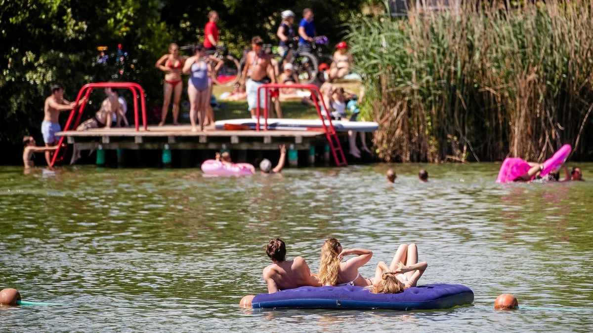 Drei Personen liegen auf einer Schwimmmatratze im Wasser, auf einem Steg stehen Menschen.