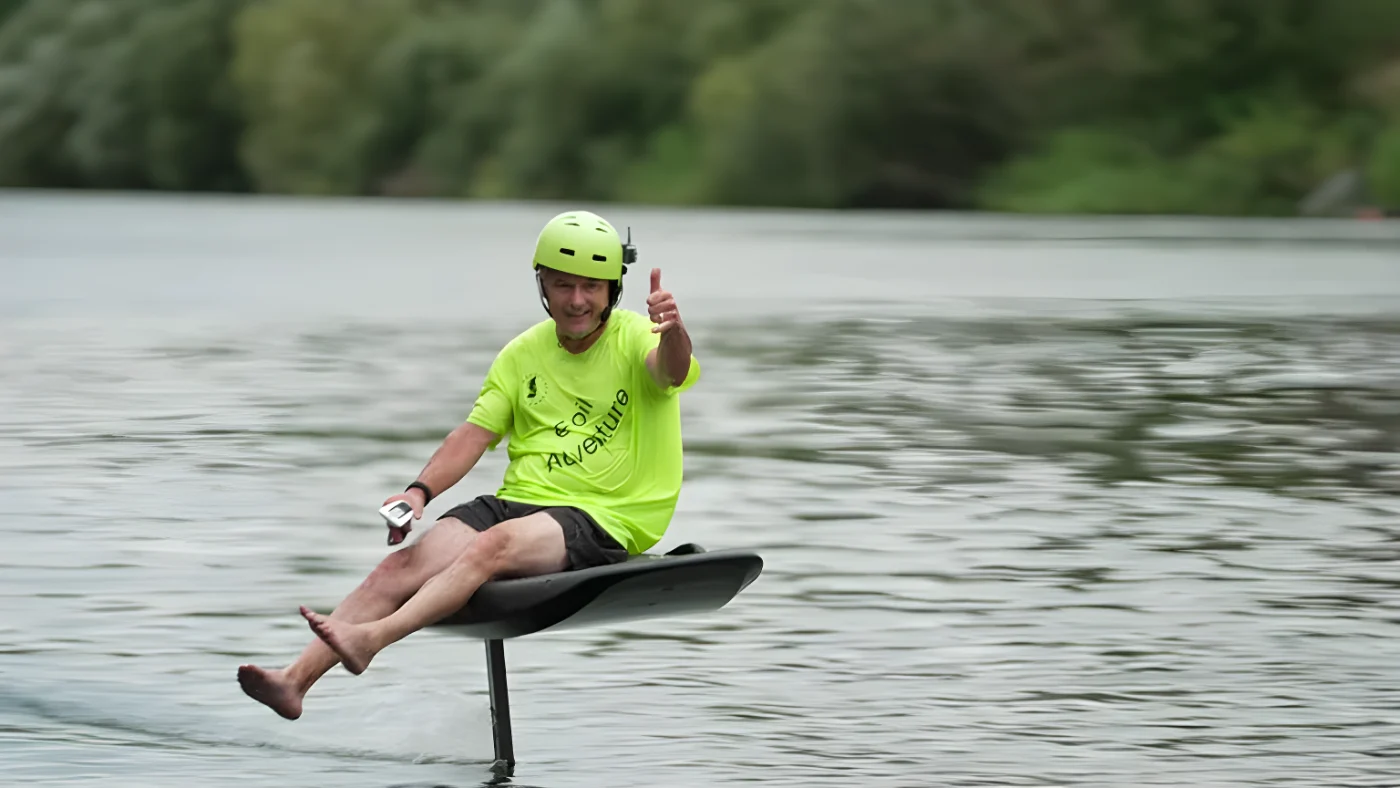Ein Mann sitzt auf einem E-Board und zeigt mit seiner Hand den Daumen hoch. Er schwebt etwa 40 Zentimeter über der Wasseroberfläche. Er trägt einen grünen Helm.
