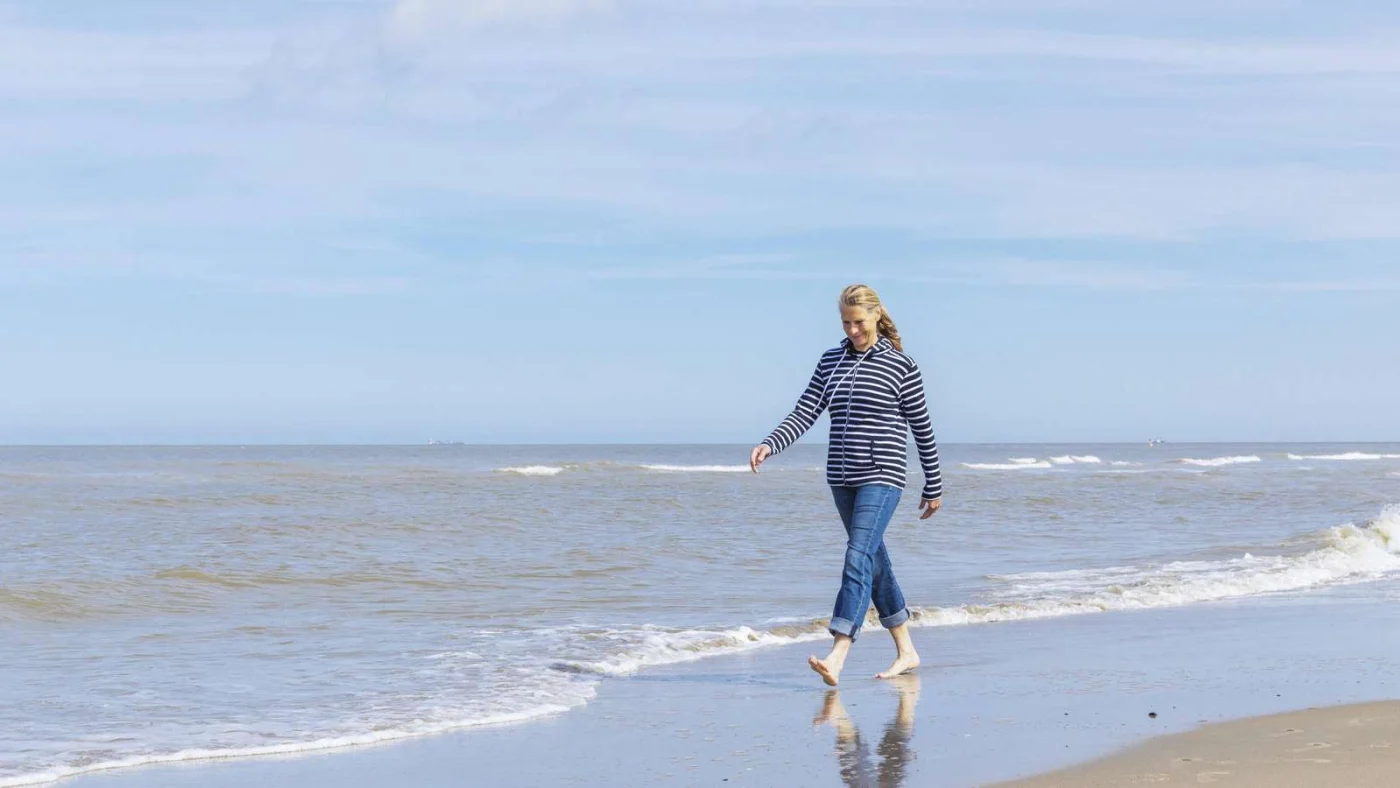 Eine lächelnde Frau mittleren Alters in Jeans und gestreiftem Hoodie läuft barfuß am Strand entlang der Wasserlinie. Der Himmel ist blau, das Meer ruhig.