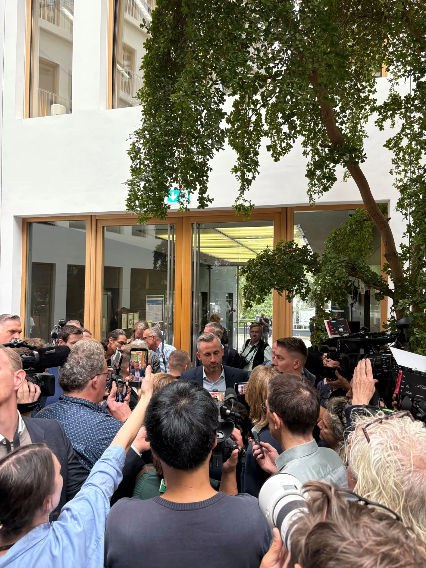 Eine Person wird von Medienvertretern mit Kameras und Mikrofonen vor einem Gebäude umringt und befragt.