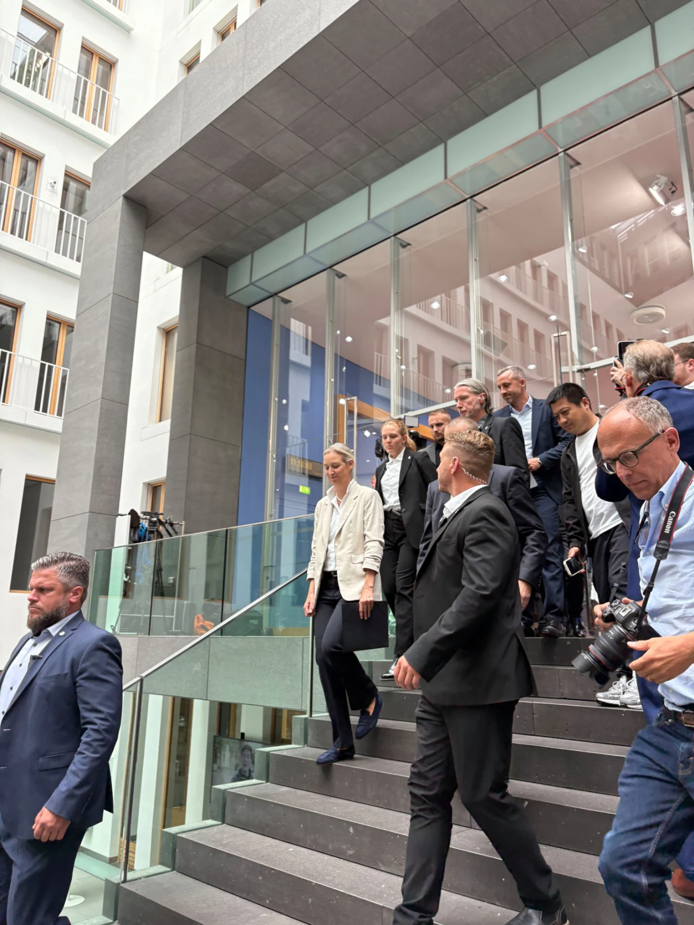 Gruppe von Personen in formeller Kleidung steigt Treppe in einem modernen Gebäude hinab, begleitet von Fotografen und Sicherheitspersonal.