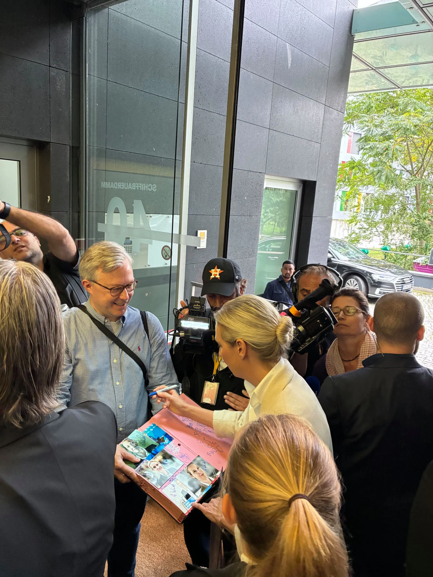Mann mit Brille und Umhängetasche hält ein Buch, umgeben von einer Menschenmenge und Kamerateams vor einem Gebäude.