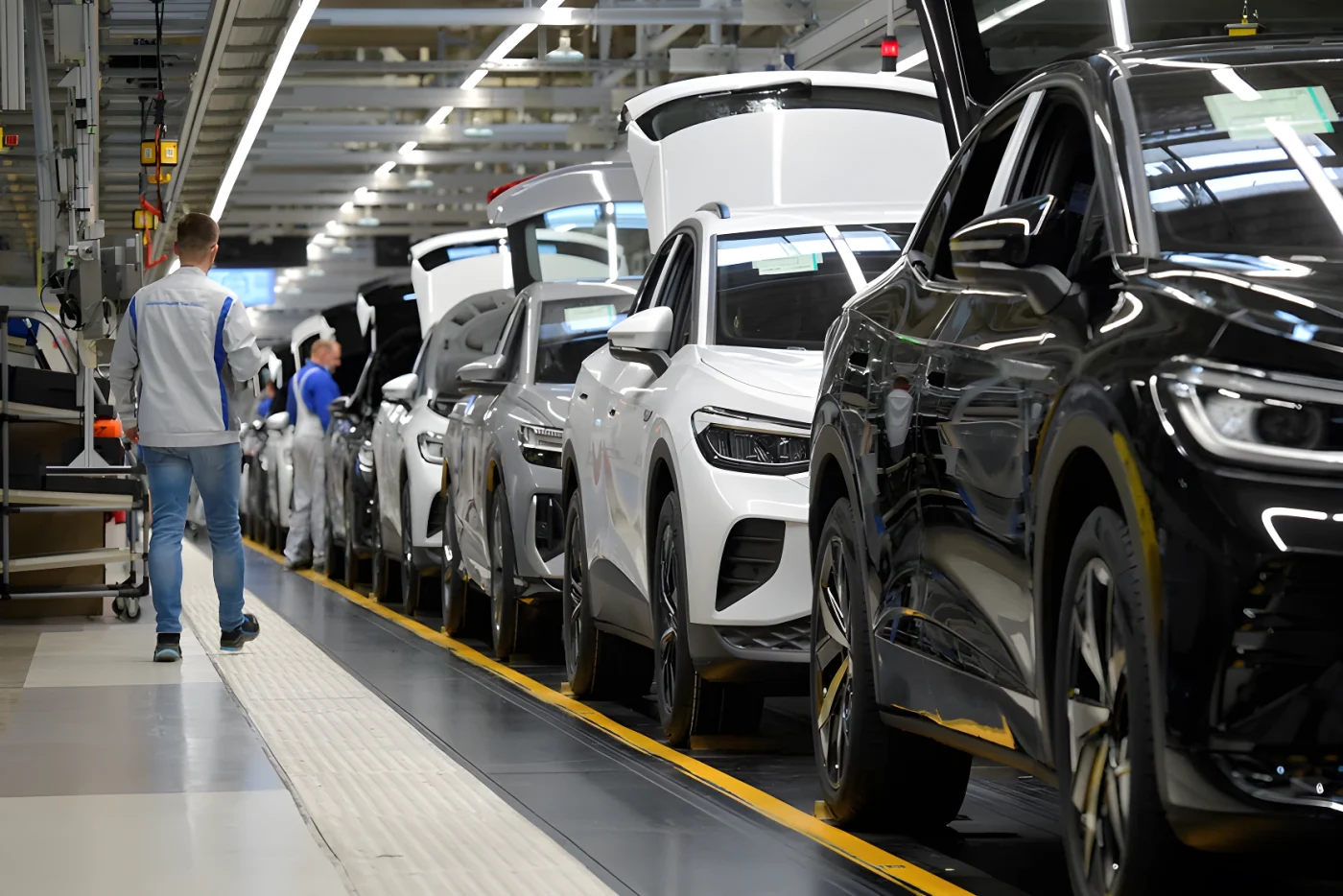 Mehrere Autos stehen hintereinander in einer Fabrik. Ein Arbeiter läuft an ihnen vorbei. Die Autos haben offene Kofferräume.