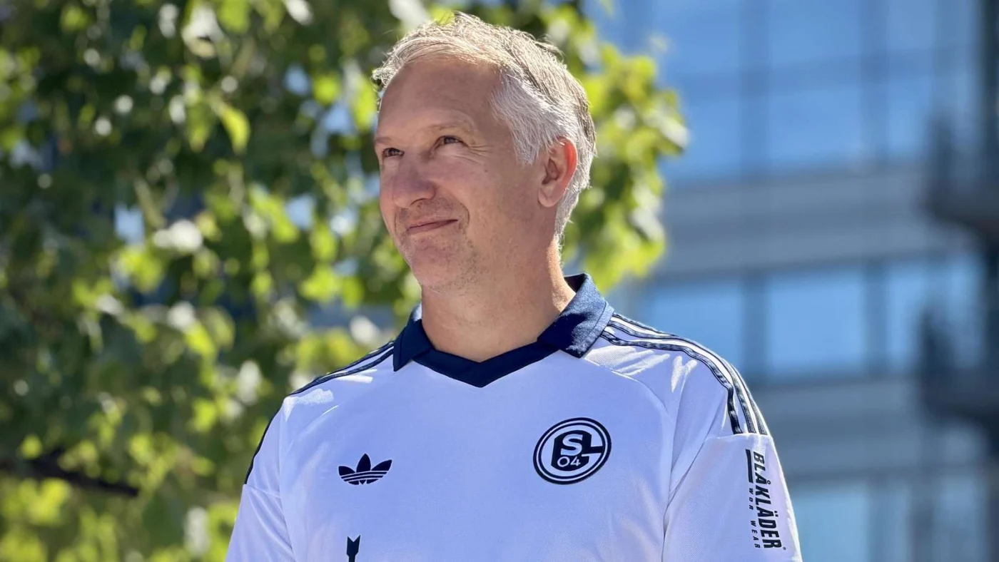 Sportvorstand Frank Baumann im Schalke-Auswärtstrikot 2026/27 lächelt.